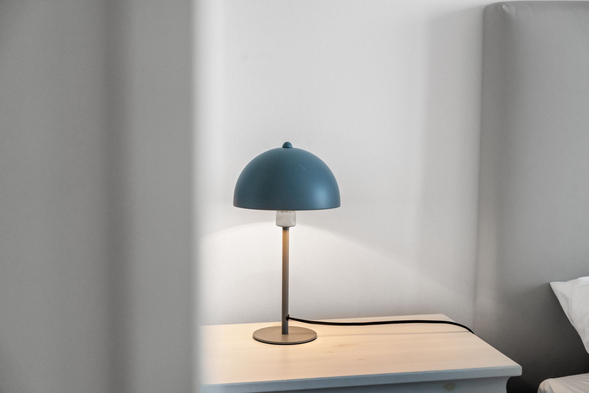 Blaue Tischlampe steht auf einem hellen Nachttisch neben einem Bett.
