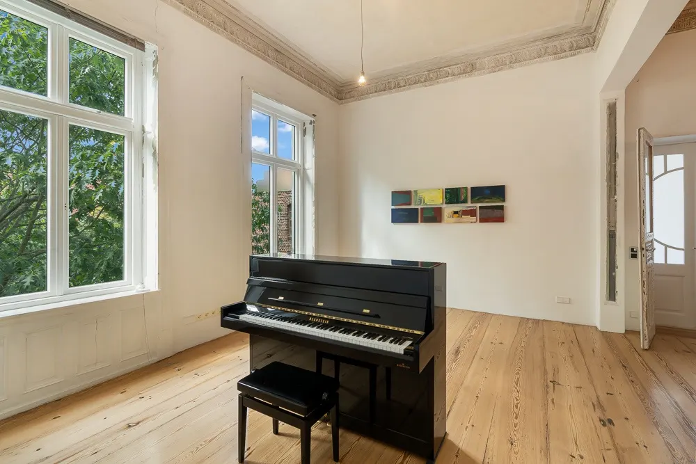 Helles Zimmer mit Holzboden, zwei großen Fenstern und einem schwarzen Klavier mit Hocker.
