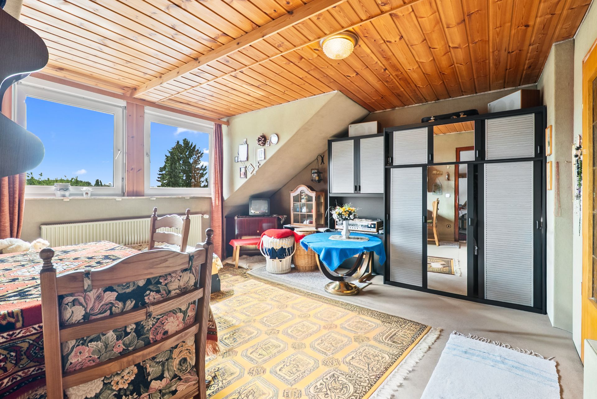 Wohnzimmer mit Holzdecke, großem Schrank, Esstisch, Teppich und zwei Fenstern mit Ausblick.