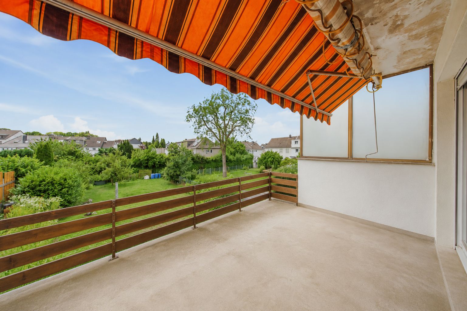 Überdachter Balkon mit orange-gestreifter Markise und Blick auf grüne Gärten und Häuser.