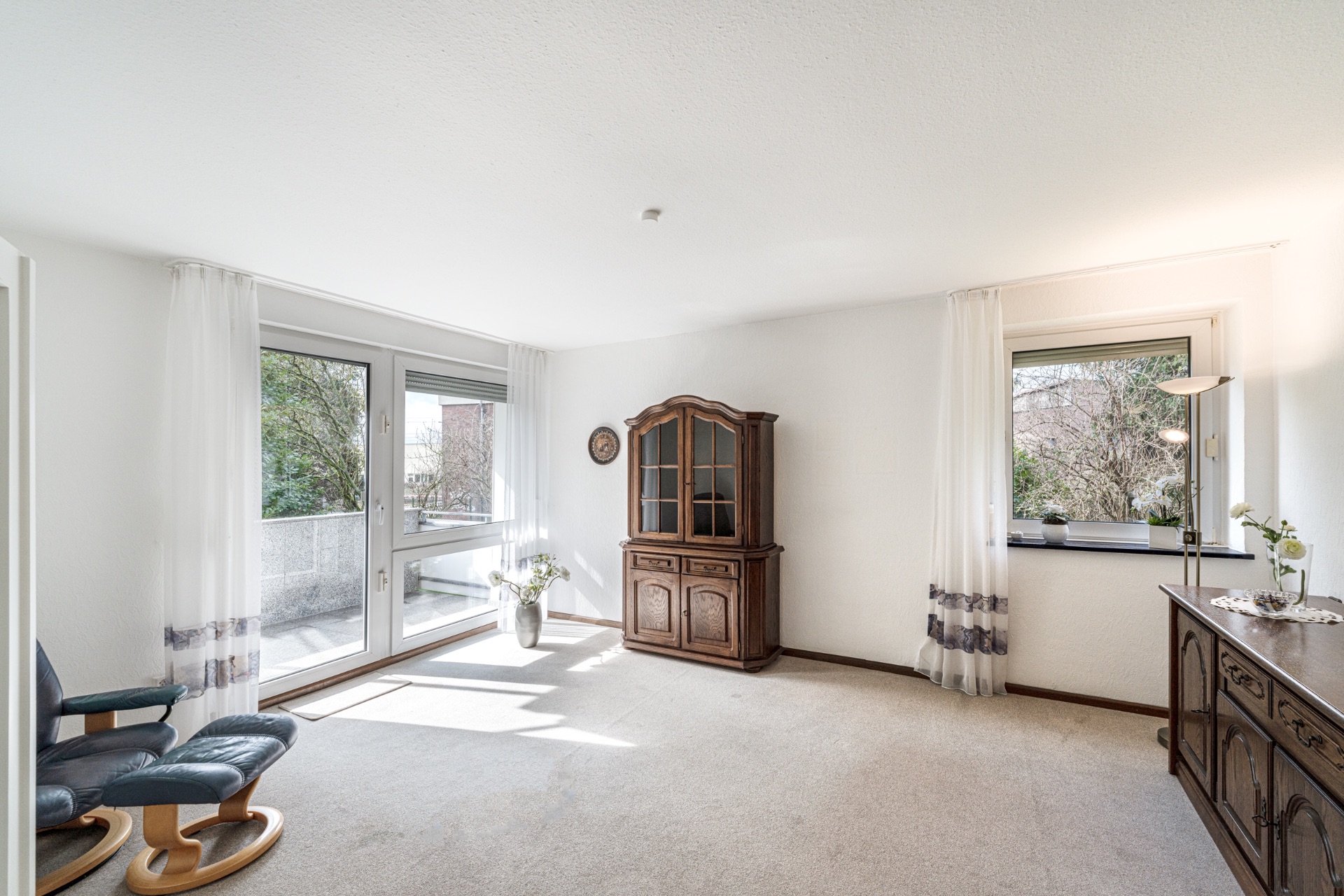 Helles Wohnzimmer mit Teppichboden, Holzvitrine, Kommode und großen Fenstern mit Blick ins Grüne.