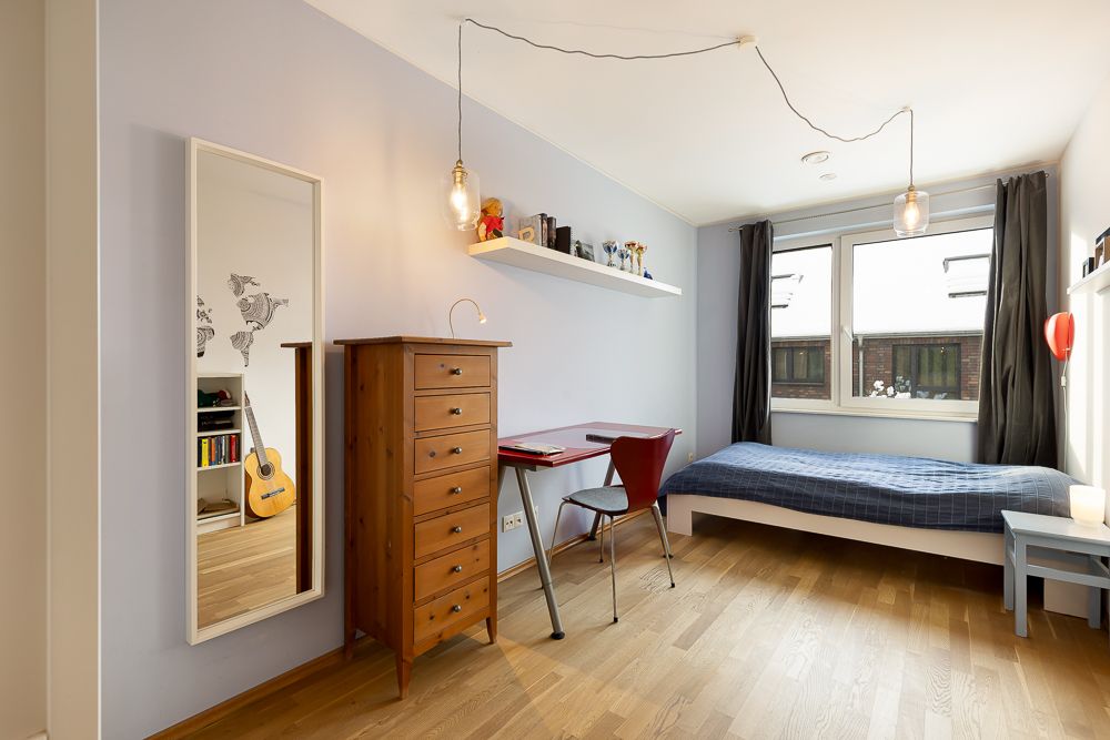 Helles Schlafzimmer mit Einzelbett, Schreibtisch, Kommode, Spiegel und großem Fenster.