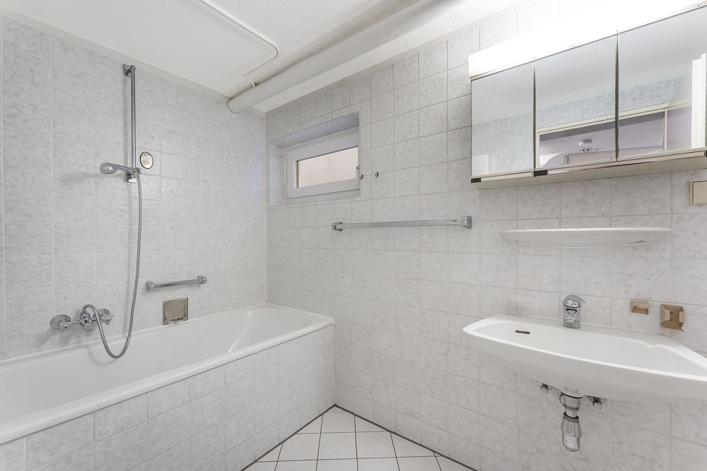 Helles Badezimmer mit Badewanne, Waschbecken und Spiegelschrank, alles in weißen Fliesen.