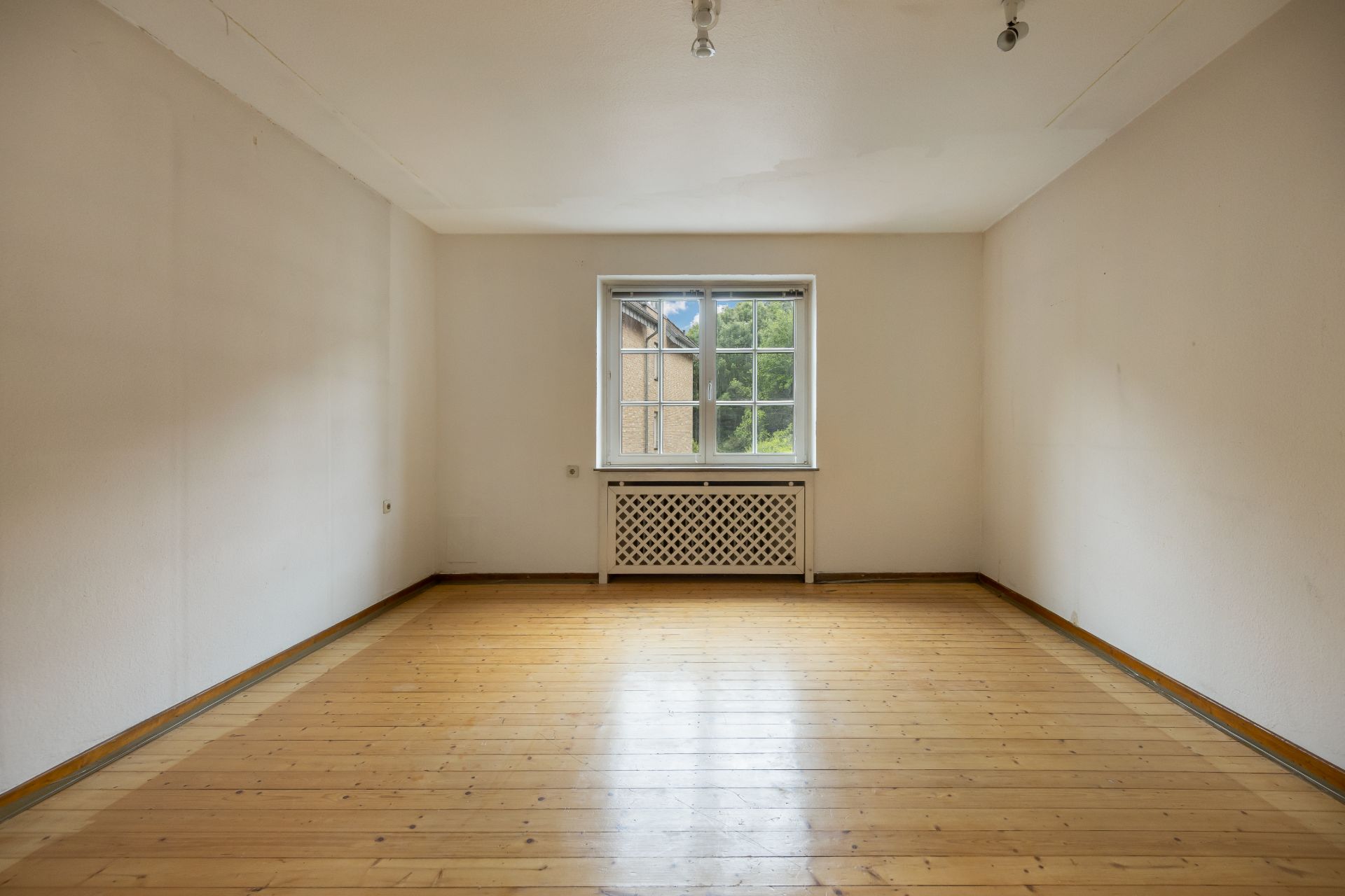 Leerer Raum mit Holzboden, weißer Heizung und Fenster mit Blick nach draußen.