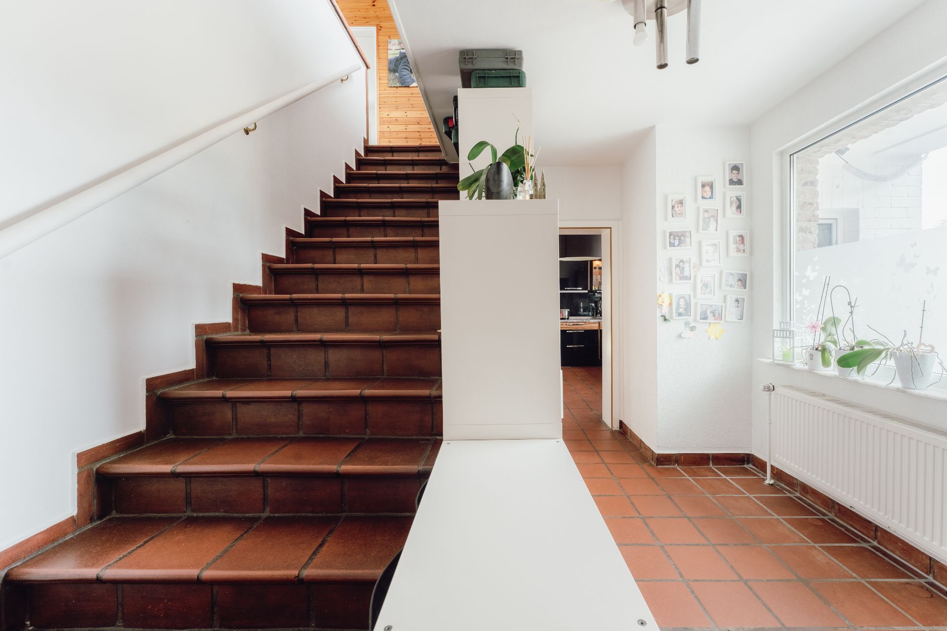 Flur mit braunen Fliesen, Treppe, weißer Wand, Pflanzen und Familienfotos an der Wand.