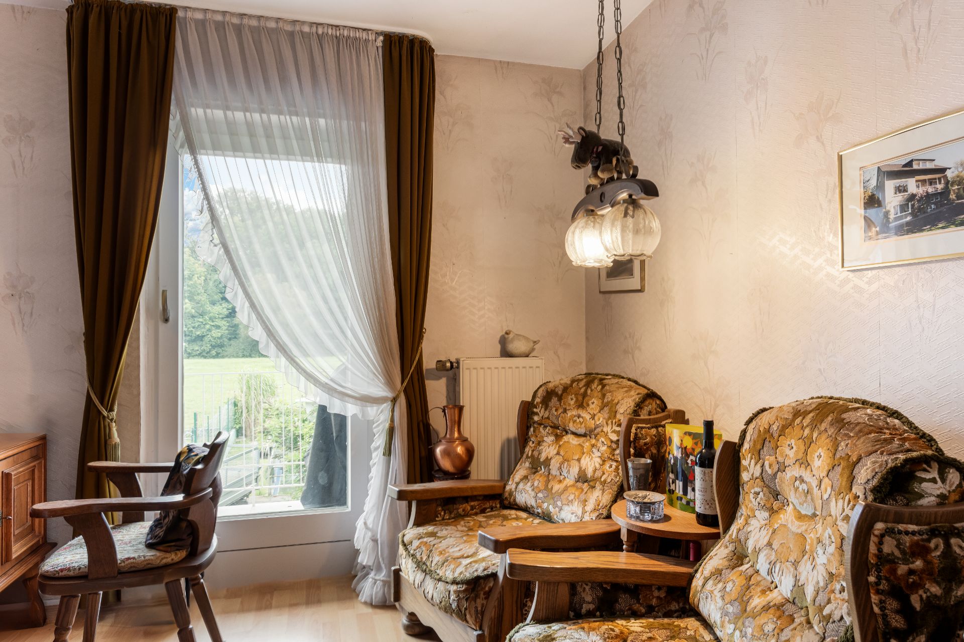 Gemütliches Wohnzimmer mit floralen Polstermöbeln, Holzstühlen, großem Fenster und braunen Vorhängen.