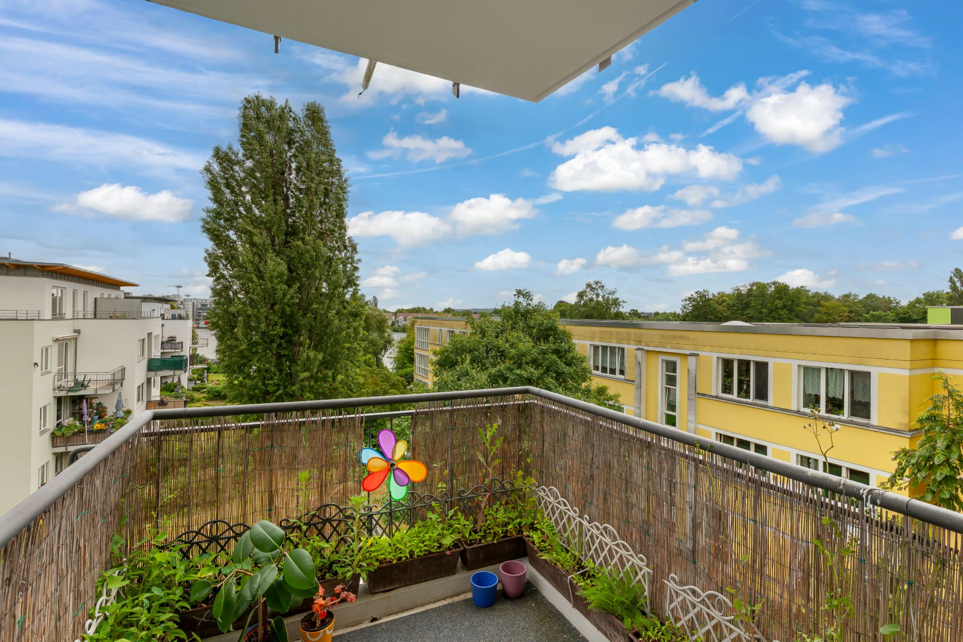 Balkon mit Pflanzen, Windrad und Ausblick auf Bäume und gelbes Wohngebäude.