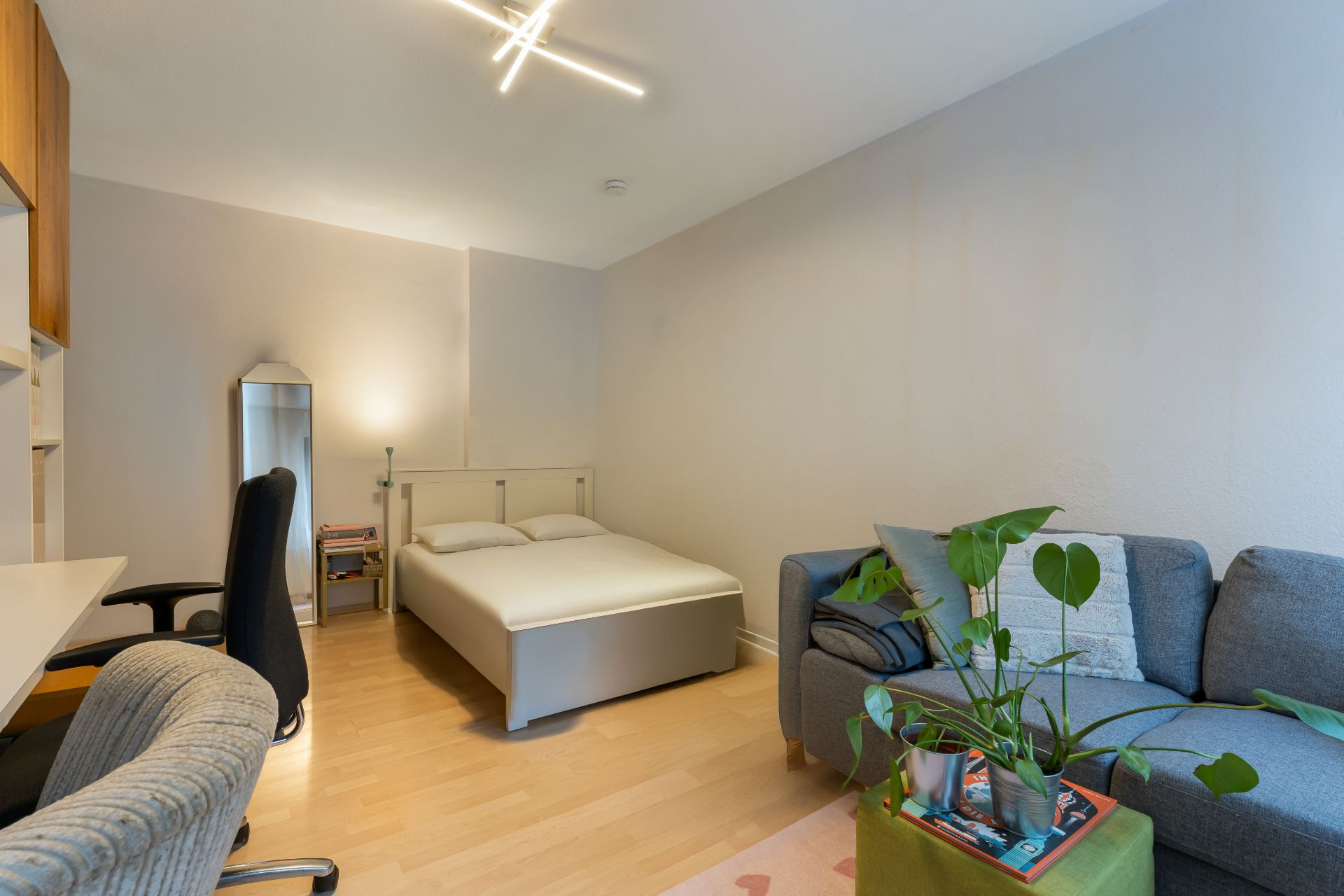 Helles Schlafzimmer mit Bett, Schreibtisch, Stuhl, Sofa und Pflanze auf hellem Holzboden.