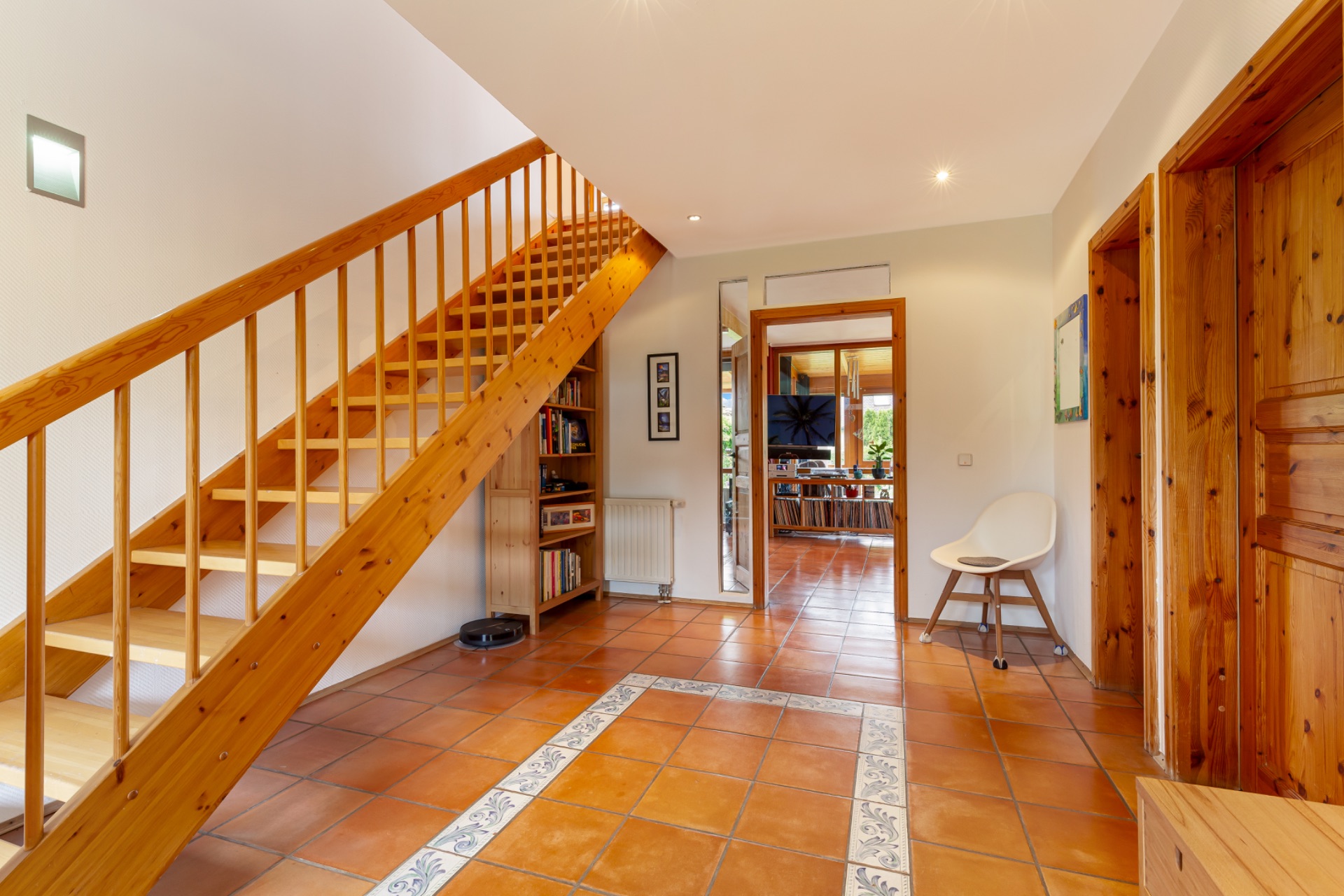 Heller Eingangsbereich mit Holztreppe, Fliesenboden, Stuhl und Bücherregal.