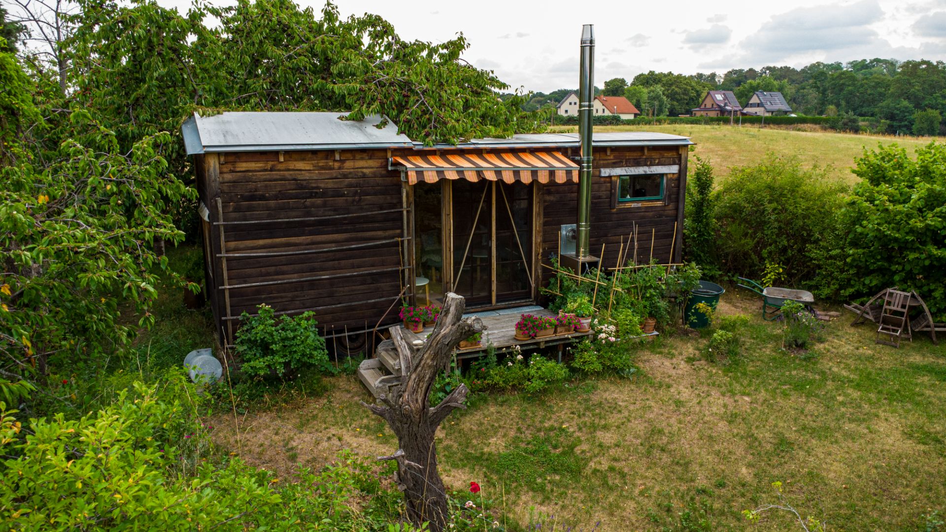 Kleines Holzhaus mit Veranda und Markise, umgeben von Garten mit Blumen und Bäumen.