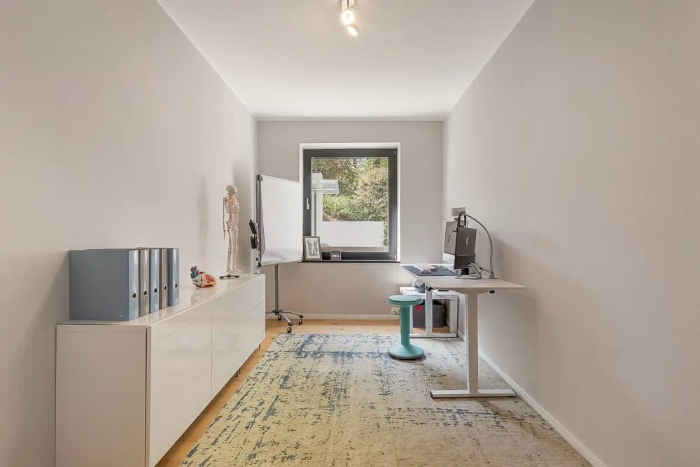 Helles Arbeitszimmer mit Schreibtisch, Stuhl, Kommode und Fenster, weiße Wände und Teppichboden.