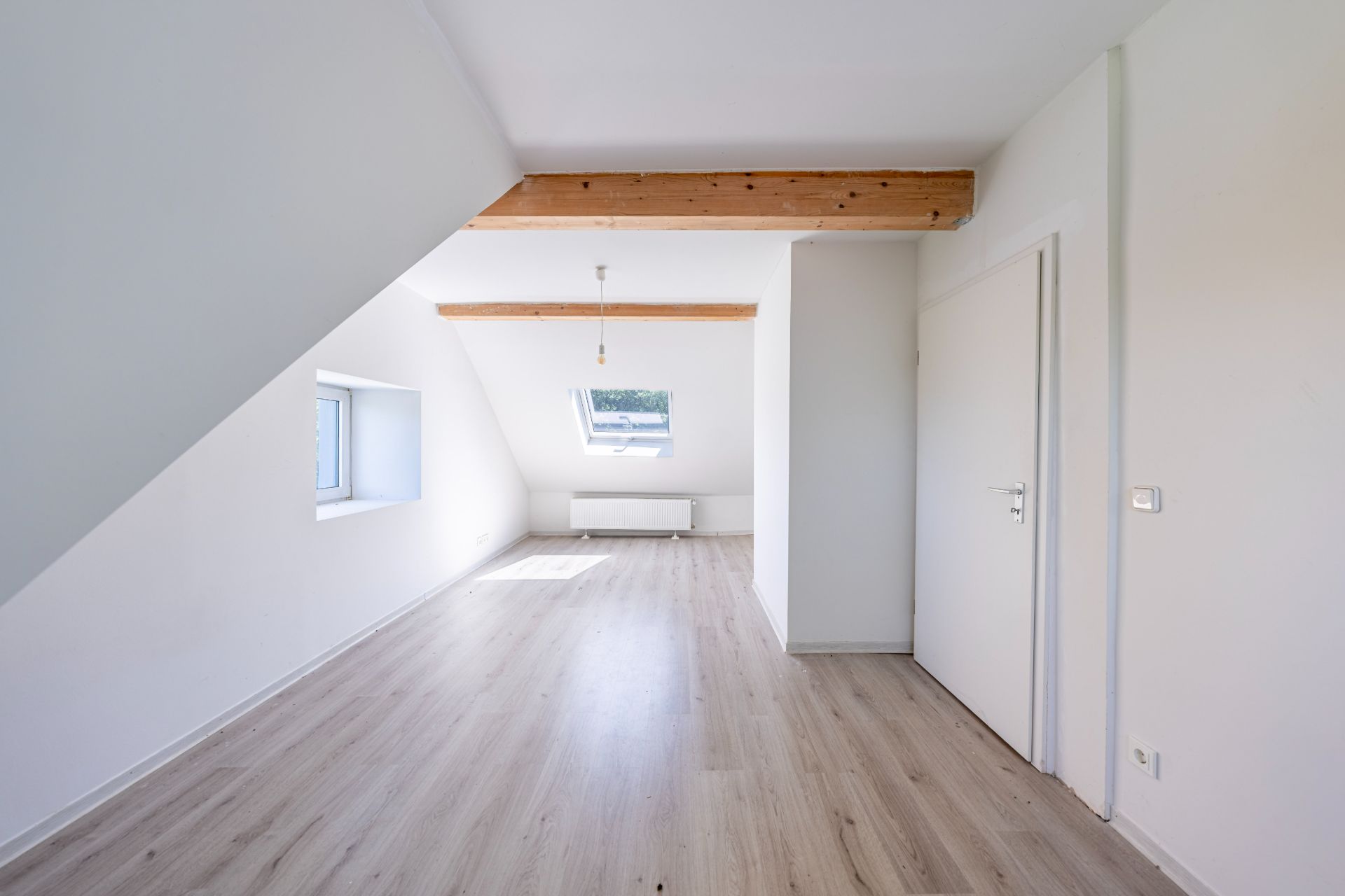 Heller, leerer Raum mit schräger Decke, zwei Fenstern und Holzbalken, heller Holzboden.