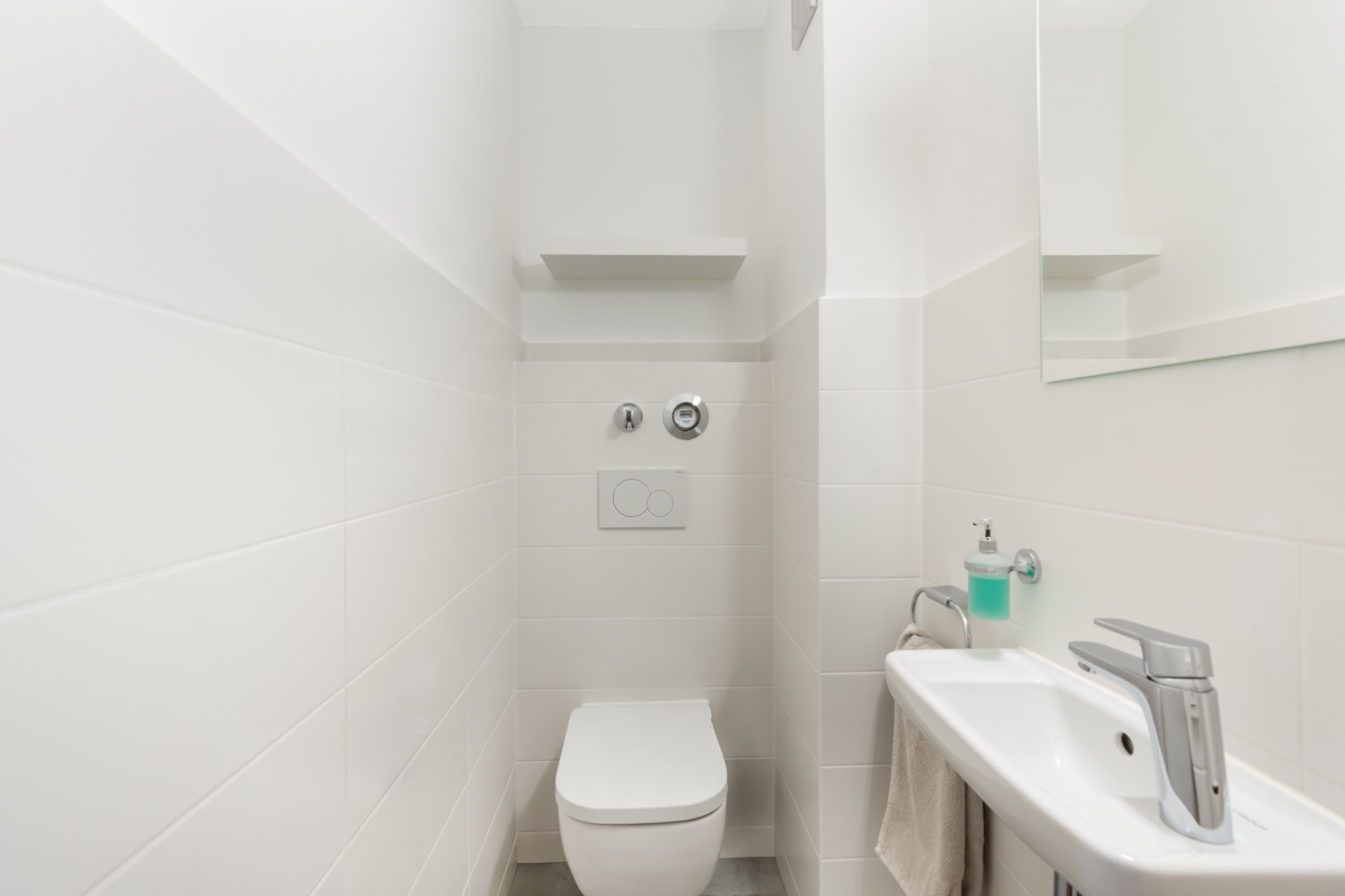 Modernes, weiß gefliestes WC mit kleinem Waschbecken und Spiegel.