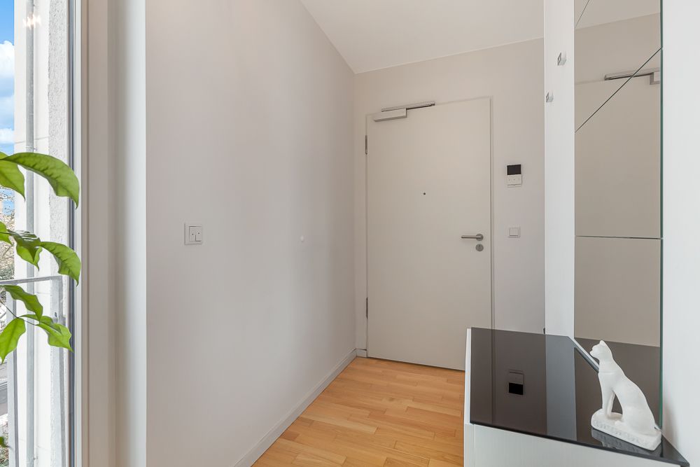 Heller Flur mit Holzboden, weißer Eingangstür und einer Pflanze am Fenster.