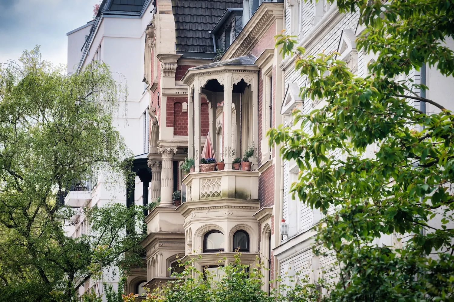 Mehrstöckiges Wohnhaus mit verzierten Balkonen und Pflanzen, umgeben von grünen Bäumen.