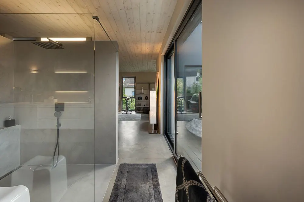 Modernes Badezimmer mit Glasdusche, großem Fenster und Blick ins Wohnzimmer.