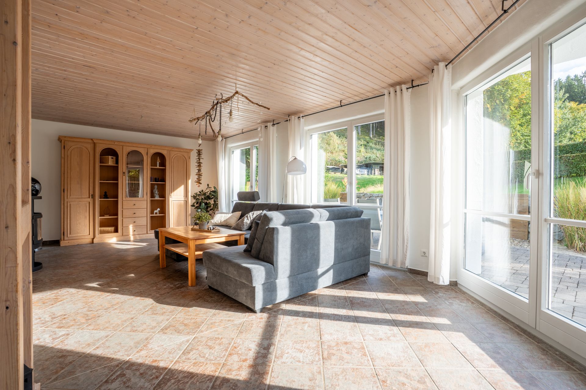 Helles Wohnzimmer mit großen Fenstern, grauem Sofa, Holztisch und Schrankwand.