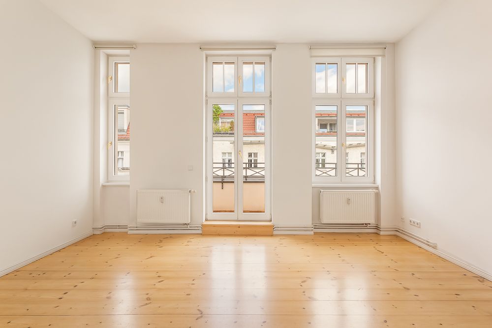Helles, leeres Zimmer mit drei großen Fenstern, Holzboden und weißen Wänden.