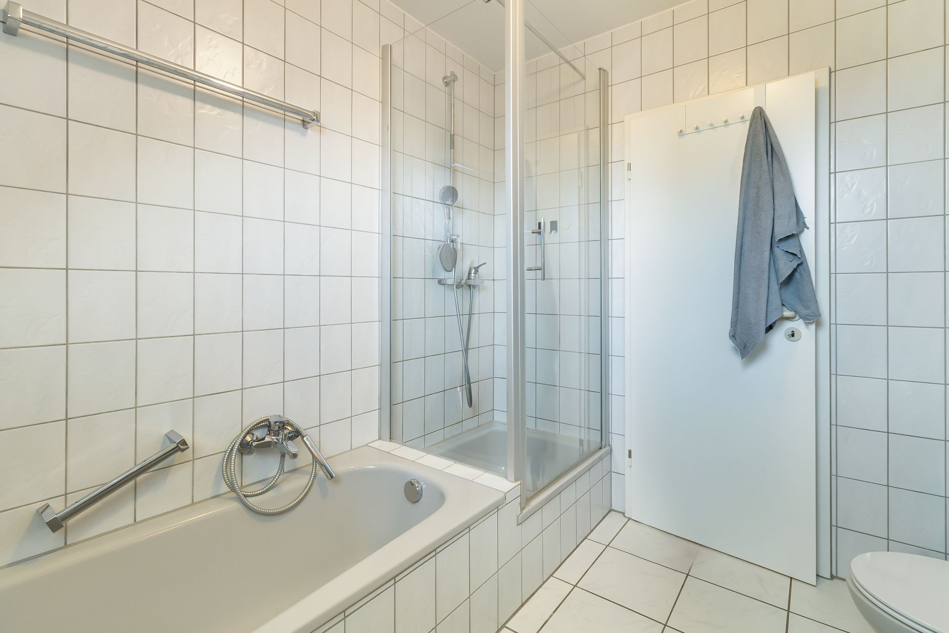 Helles Badezimmer mit Badewanne, Dusche und Handtuch an einer weißen Tür.