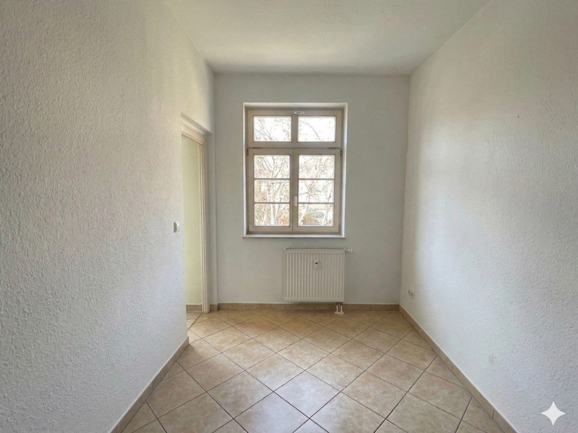 Leerer, heller Raum mit Fliesenboden, Fenster und Heizkörper an der Wand.