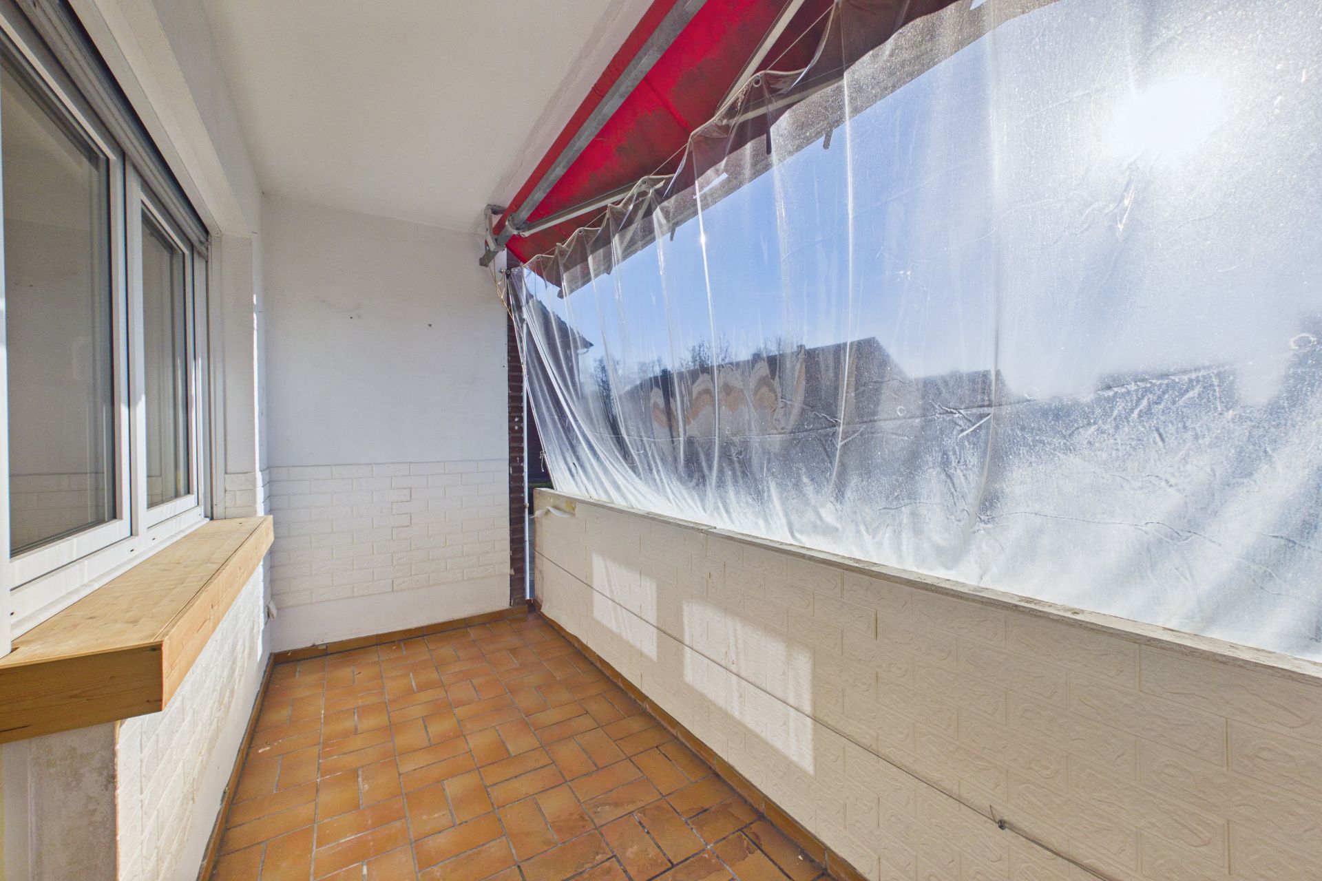 Überdachter Balkon mit Fenster, Fliesenboden und durchsichtiger Plane als Sichtschutz.