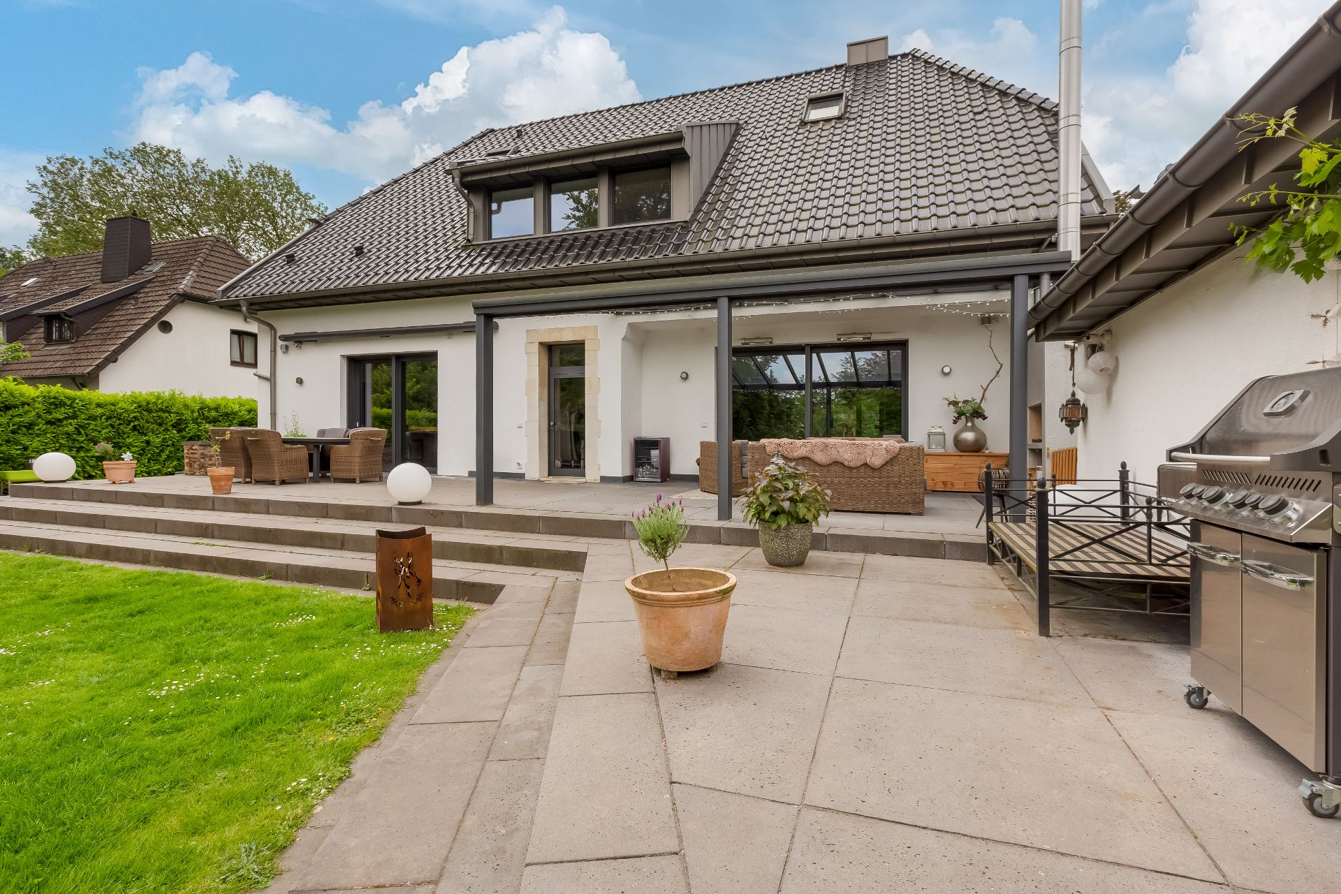 Große Terrasse mit Sitzmöbeln, Grill und bepflanzten Töpfen vor einem Einfamilienhaus.