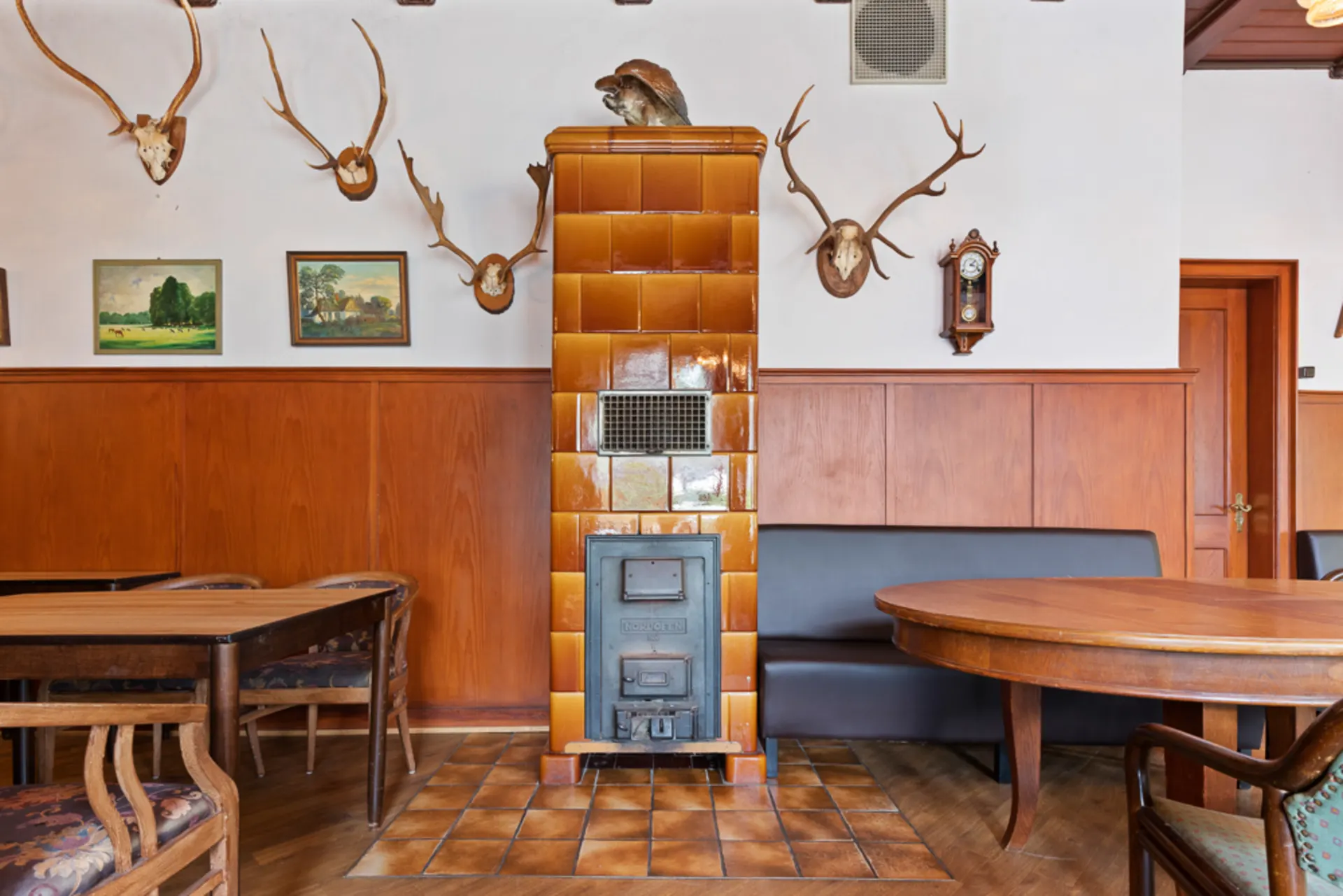 Wohnzimmer mit Kachelofen, Holzmöbeln, Geweihen und Bildern an der Wand.