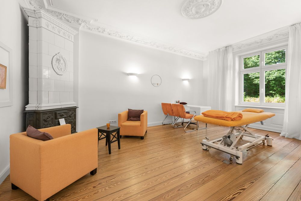 Heller Raum mit zwei orangefarbenen Sesseln, Behandlungsliege, Schreibtisch und großem Fenster.