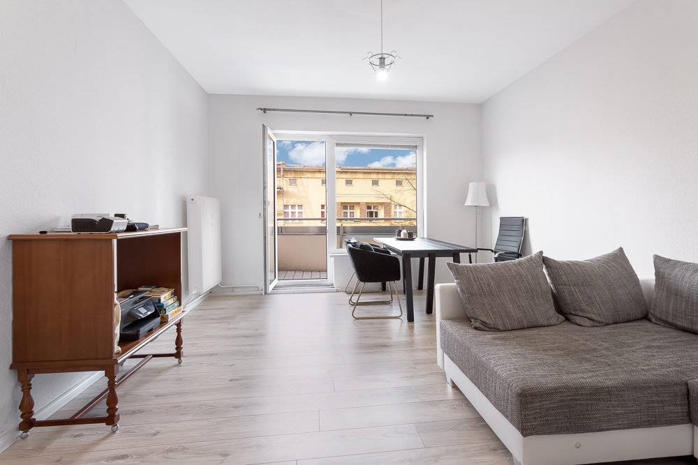 Helles Wohnzimmer mit Sofa, Esstisch, Stühlen, Regal und Zugang zu einem Balkon.