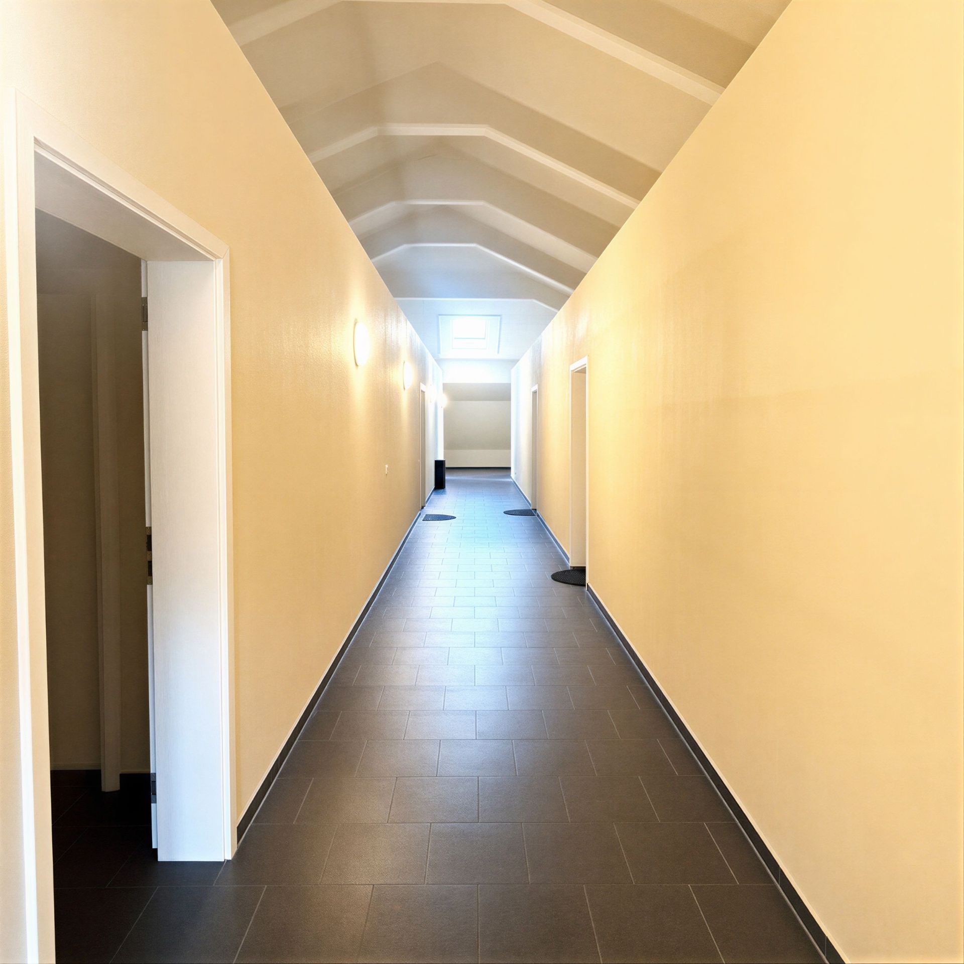Heller Flur mit mehreren Türen und Dachfenster, dunkler Fliesenboden.