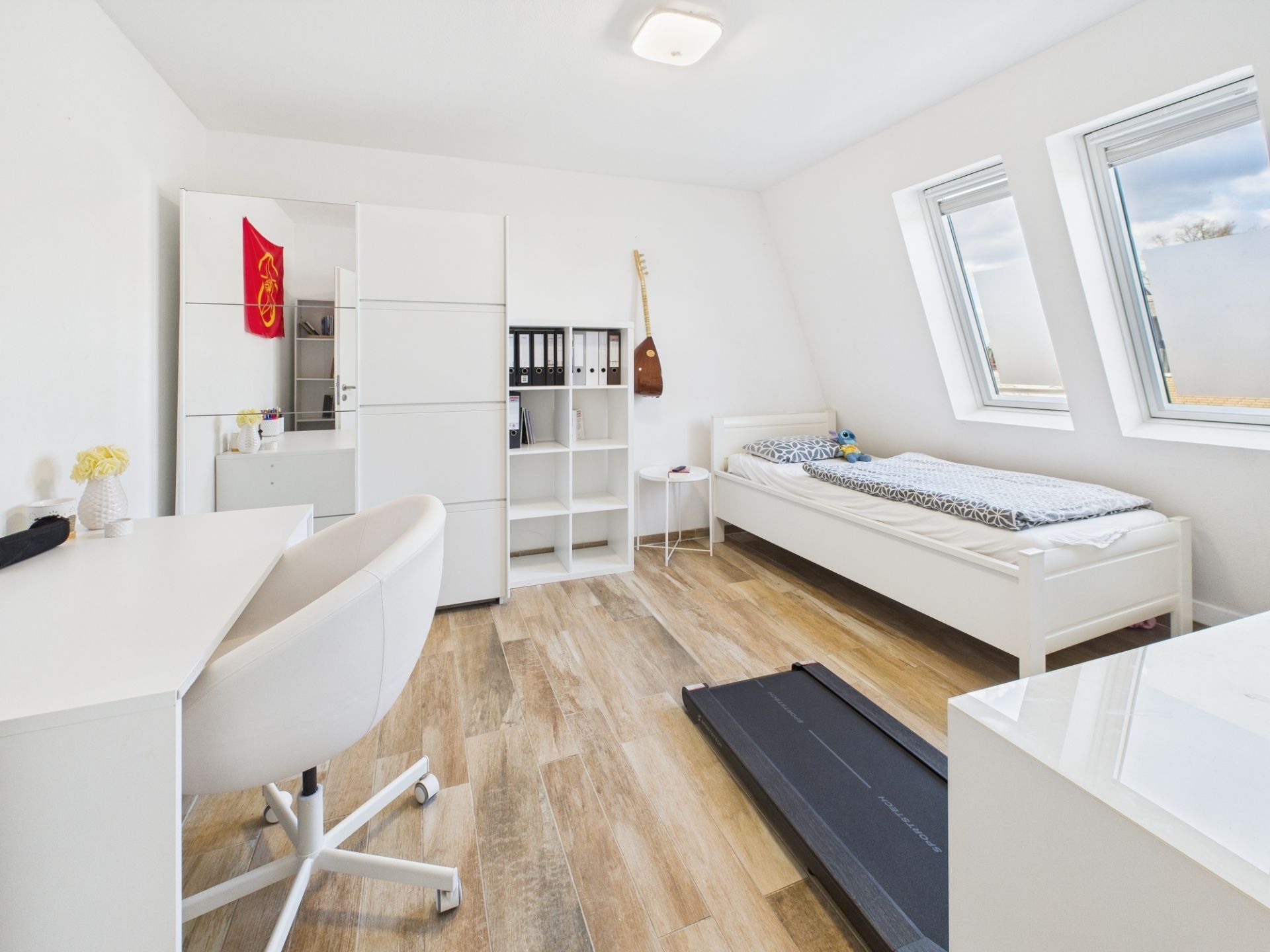 Helles Zimmer mit Einzelbett, Schreibtisch, Regal, Schrank, Laufband und zwei Fenstern.