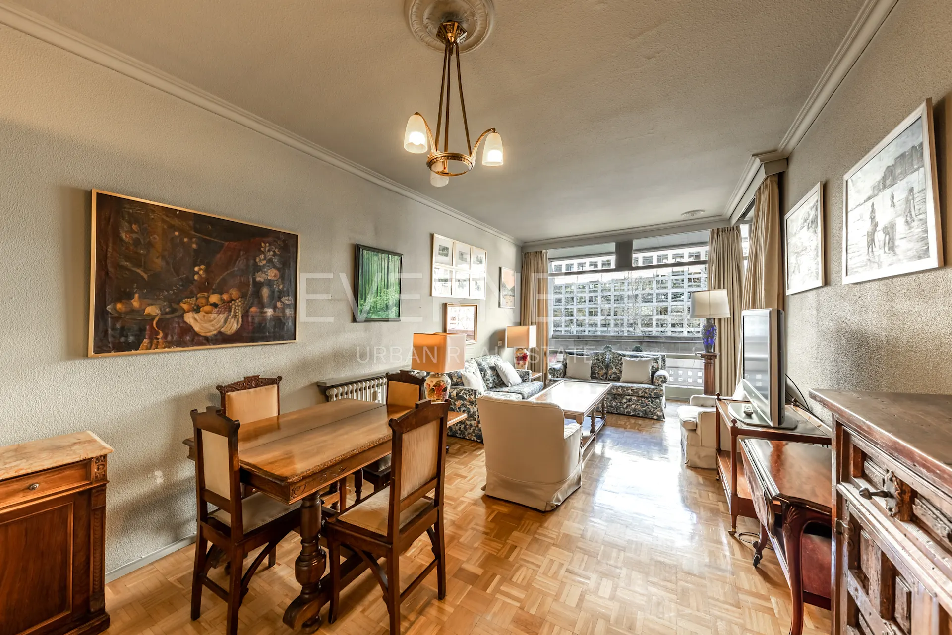 Helles Wohnzimmer mit Holztisch, Sesseln, Sofa, Fernseher und mehreren Bildern an den Wänden.