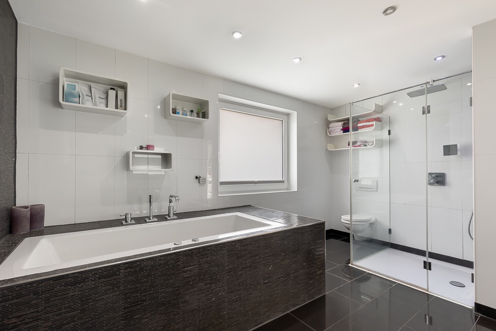 Modernes Badezimmer mit großer Badewanne, Glasdusche, Regal und Fenster.