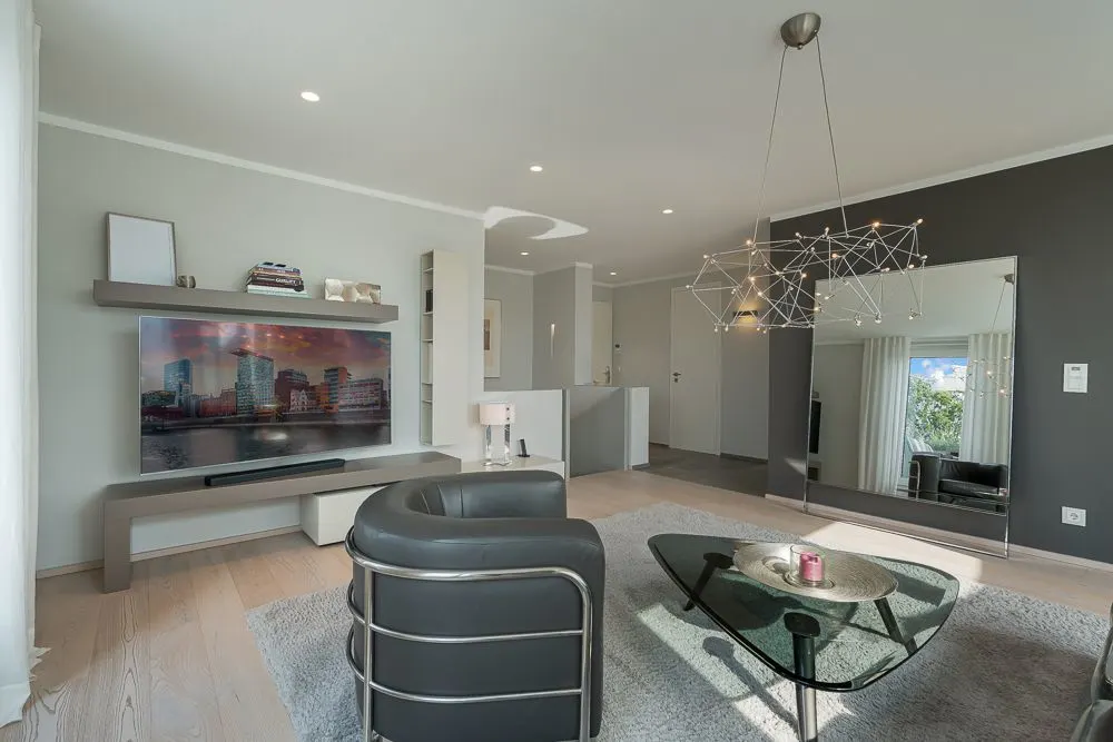 Modernes Wohnzimmer mit Sessel, großem Fernseher, Glastisch und Wandspiegel.
