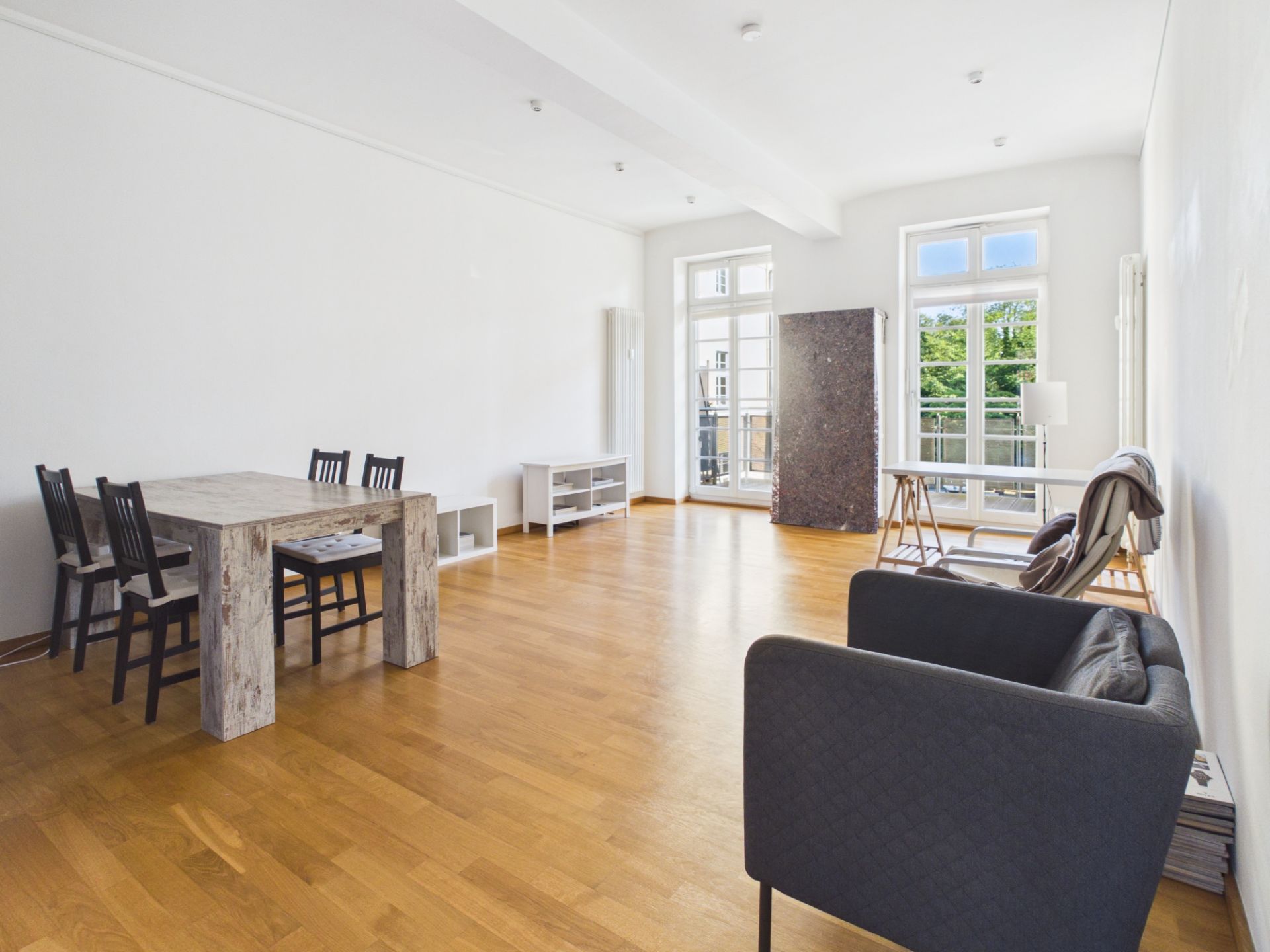 Helles Wohnzimmer mit Holzboden, Esstisch, Stühlen, Sofa und großen Fenstern.
