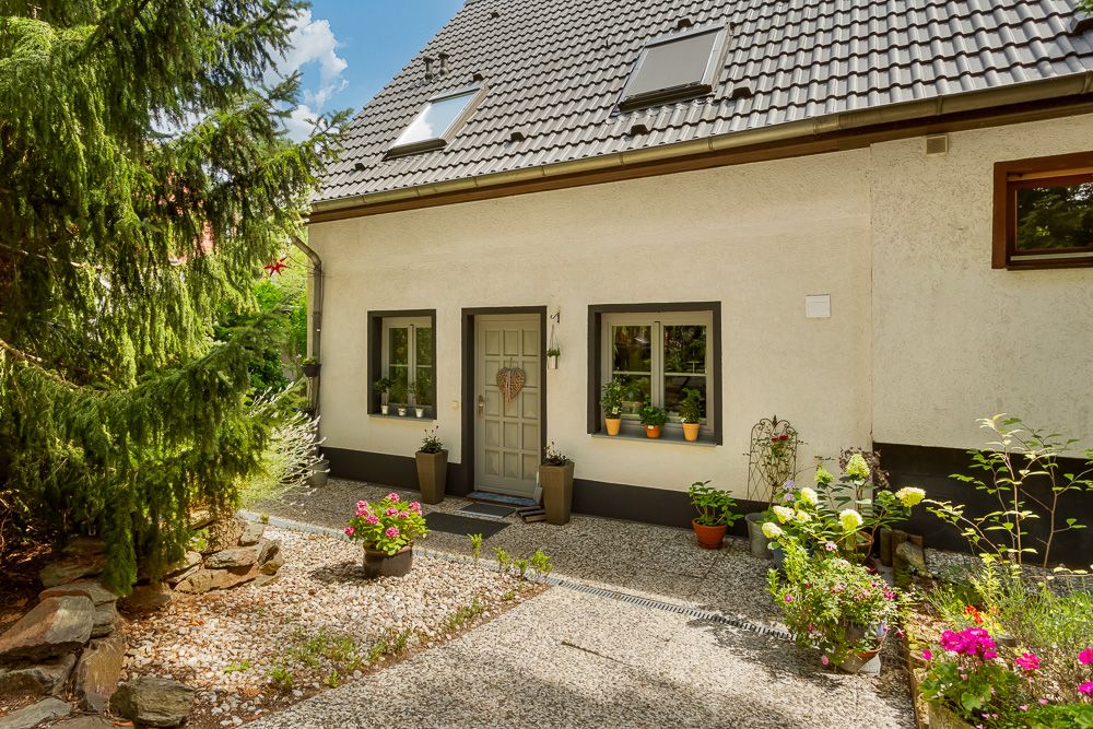 Eingangsbereich eines Hauses mit zwei Fenstern, Blumen und Topfpflanzen vor der Tür.