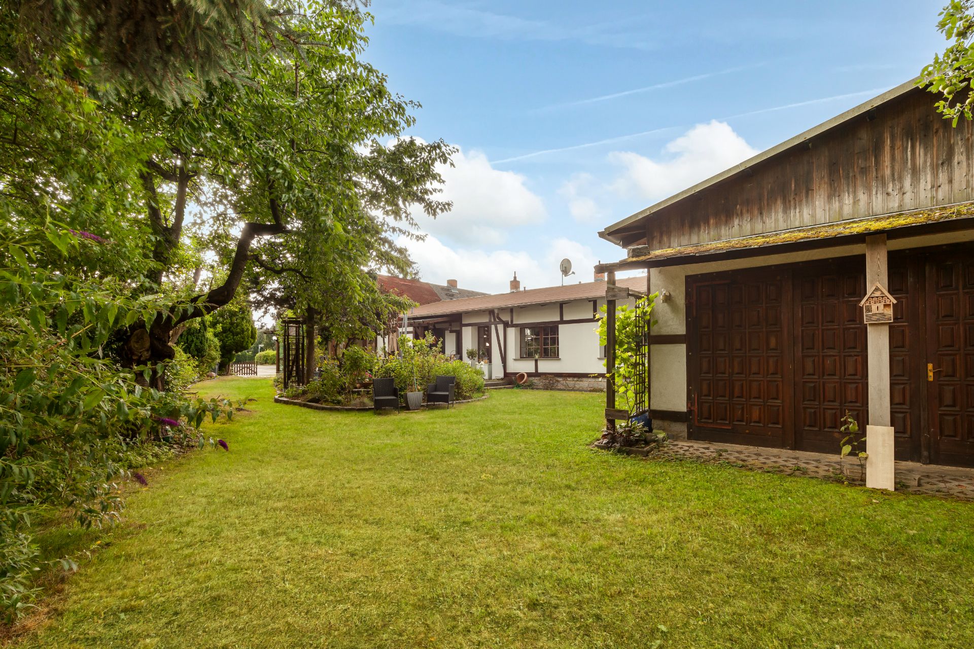 Grüner Garten mit Bäumen, Rasen, Sitzbereich und Wohnhaus im Hintergrund.