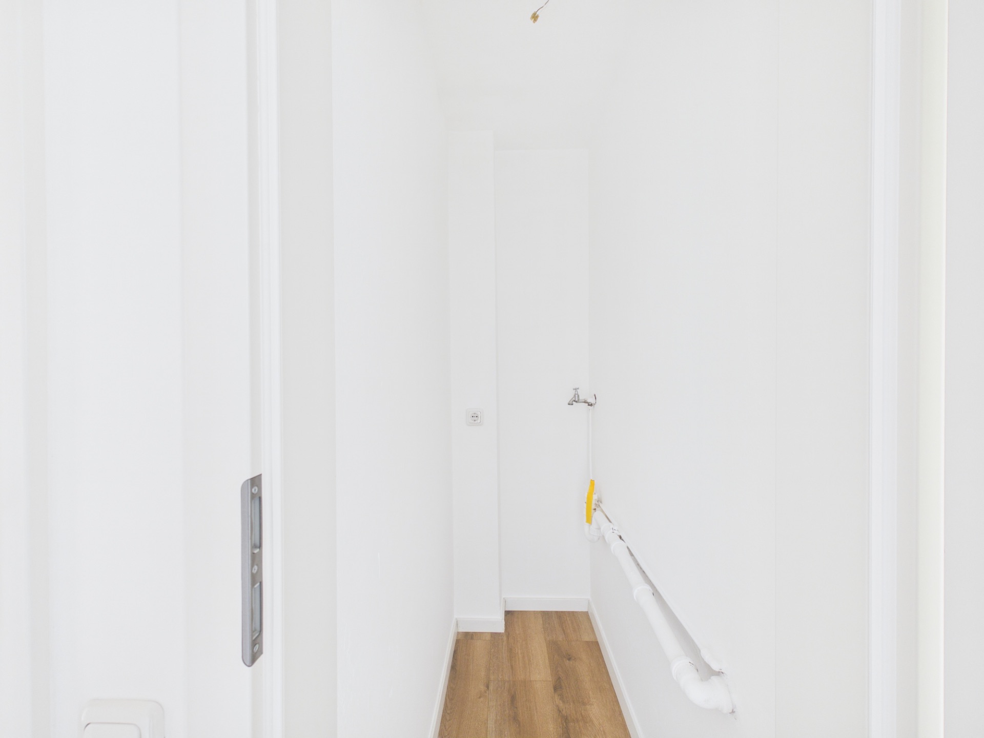 Schmaler, weißer Flur mit Holzfußboden und sichtbaren Rohren an der rechten Wand.