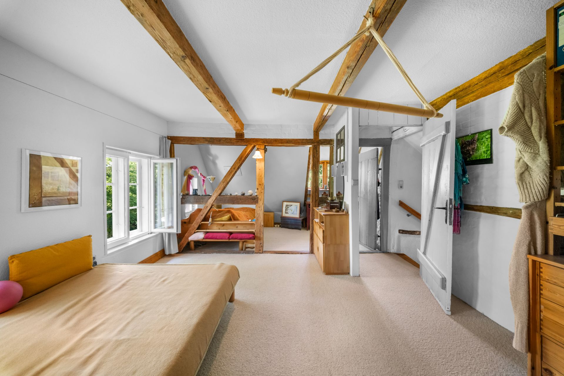 Helles Schlafzimmer mit sichtbaren Holzbalken, Doppelbett und Fenster, Teppichboden und Holzmöbeln.