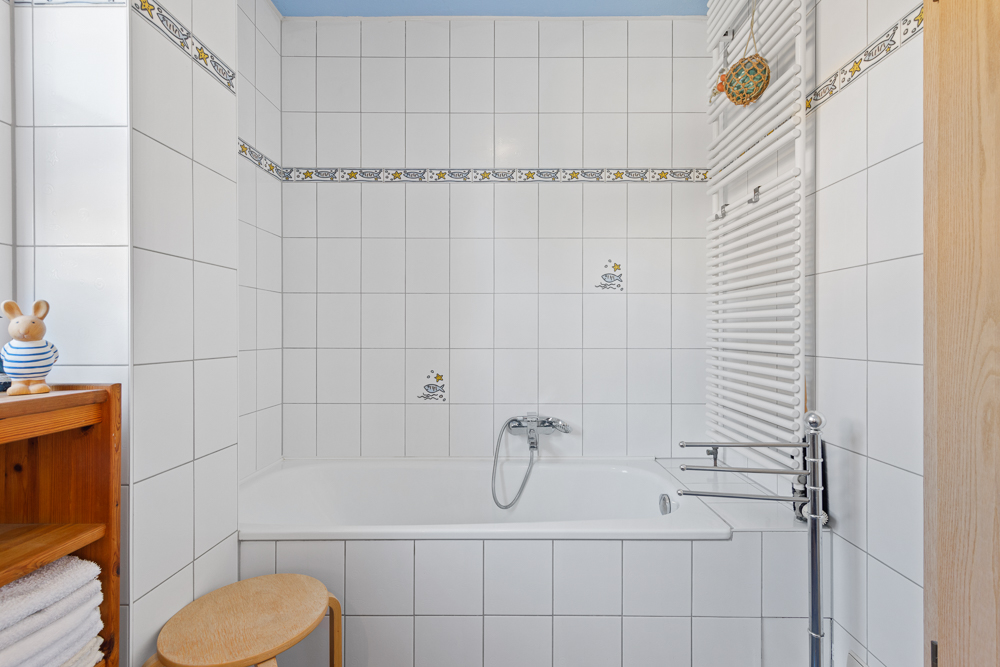Weiß gefliestes Badezimmer mit Badewanne, Heizkörper, Holzregal und Hocker.