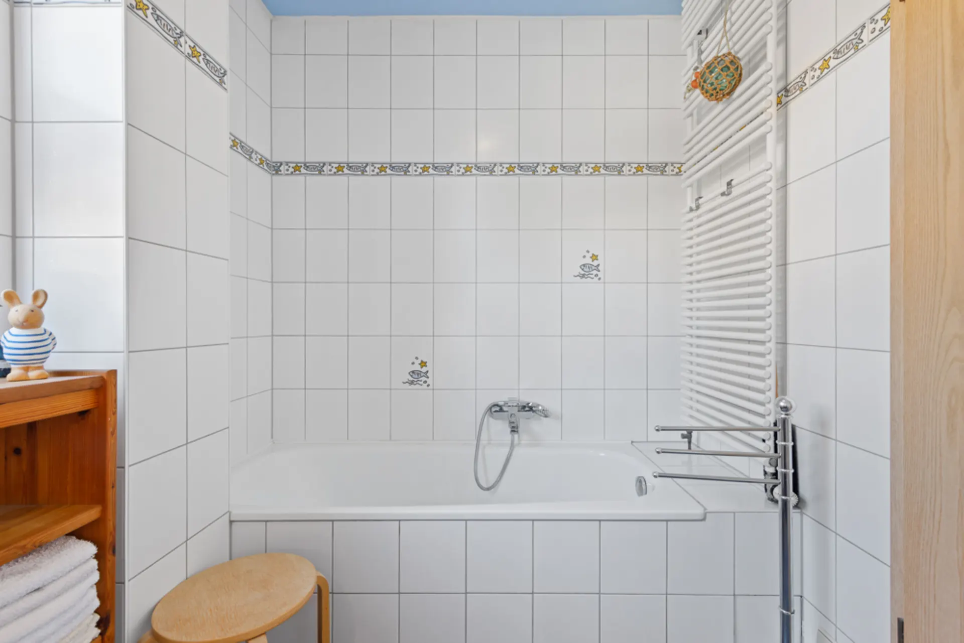 Weiß gefliestes Badezimmer mit Badewanne, Heizkörper, Holzregal und Hocker.