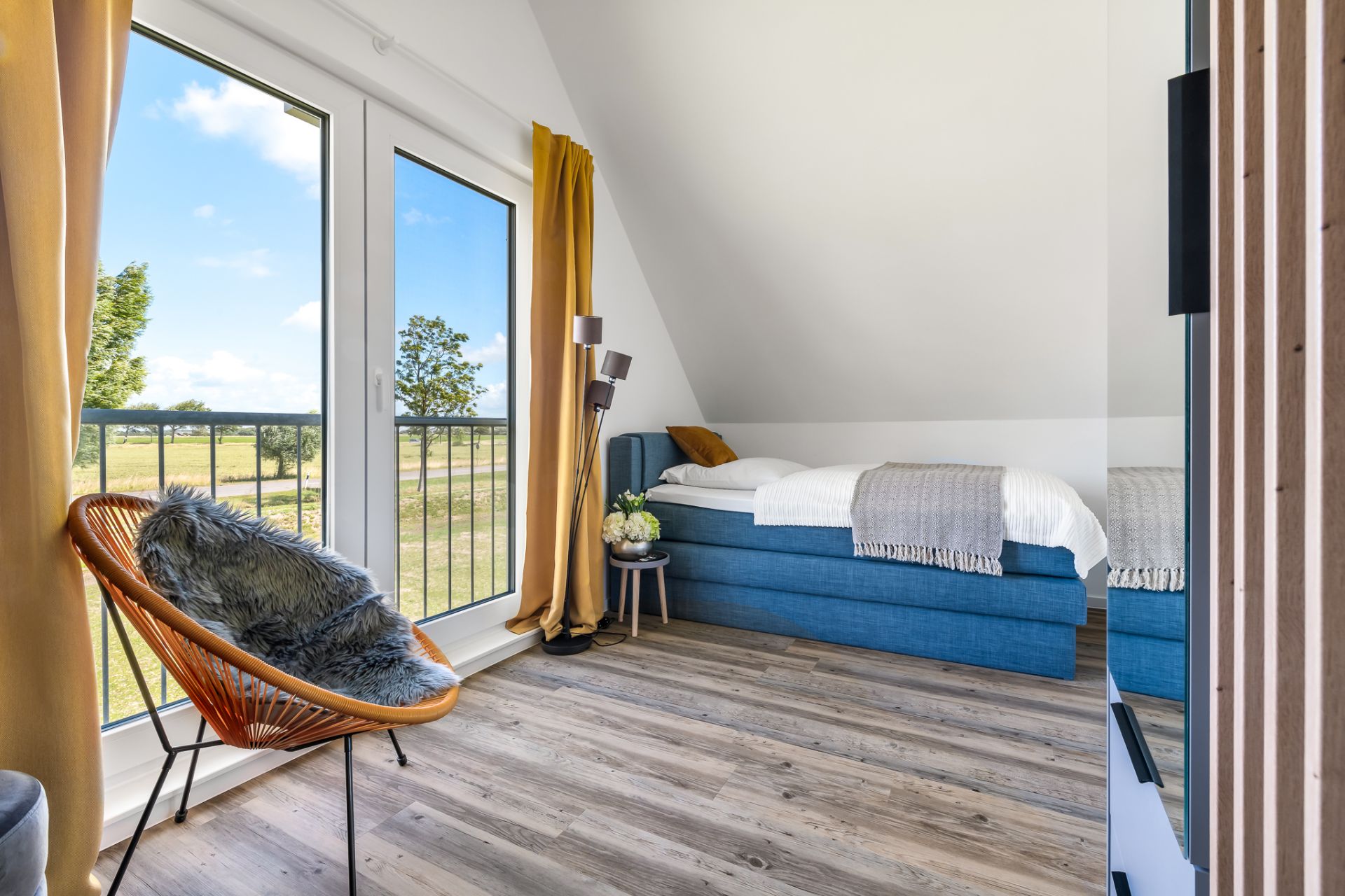 Helles Schlafzimmer mit Einzelbett, großem Fenster, gelben Vorhängen und Sessel mit Fellkissen.