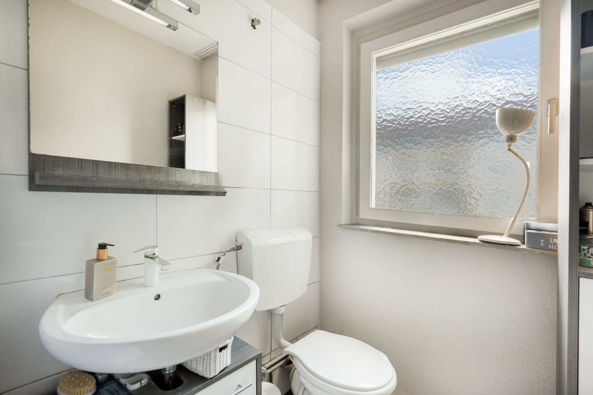 Kleines Badezimmer mit Waschbecken, Spiegel, Toilette und Fenster mit Milchglas.