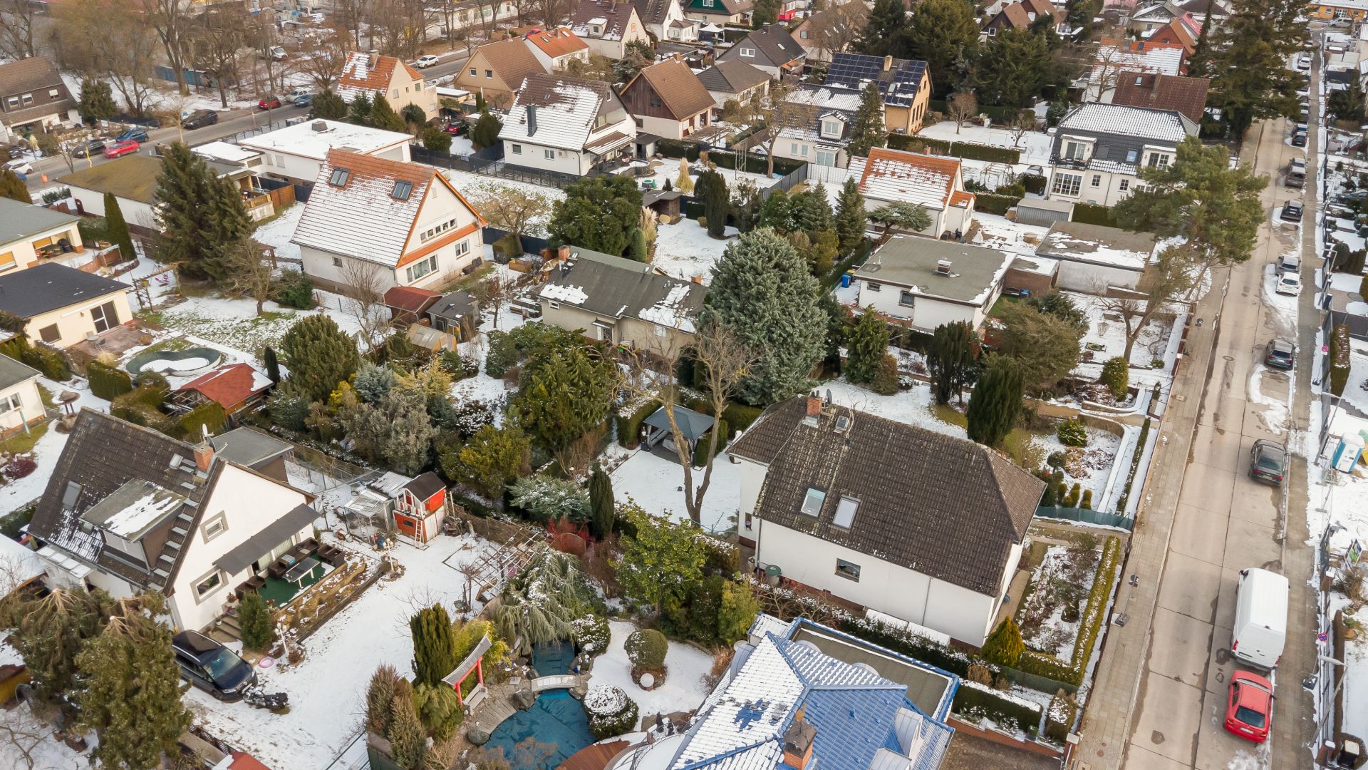 Luftaufnahme eines Wohngebiets mit Häusern, Gärten und schneebedeckten Dächern im Winter.