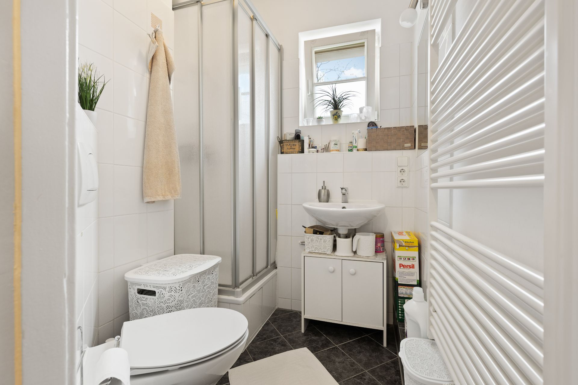 Kleines Badezimmer mit Dusche, Waschbecken, Toilette, Fenster und Handtuchheizkörper.