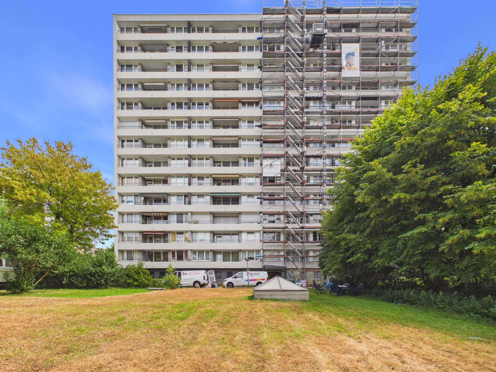Mehrstöckiges Wohnhaus mit Baugerüst an einer Seite, davor grüne Wiese und Bäume.
