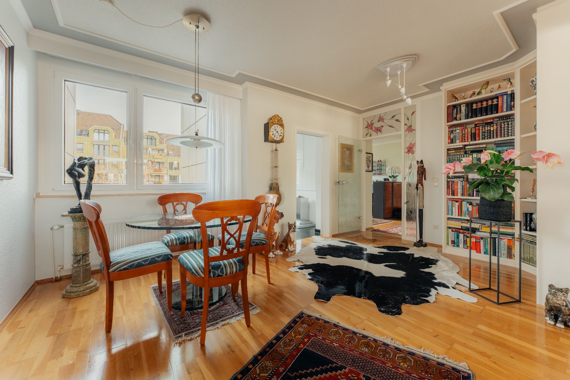 Helles Wohnzimmer mit Esstisch, Holzstühlen, Bücherregal, Teppichen und großen Fenstern.