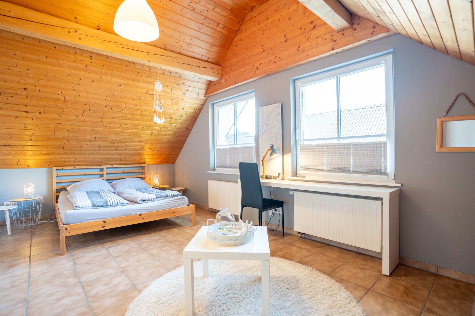 Helles Schlafzimmer mit Holzdach, Bett, Schreibtisch, Stuhl und großem Fenster.
