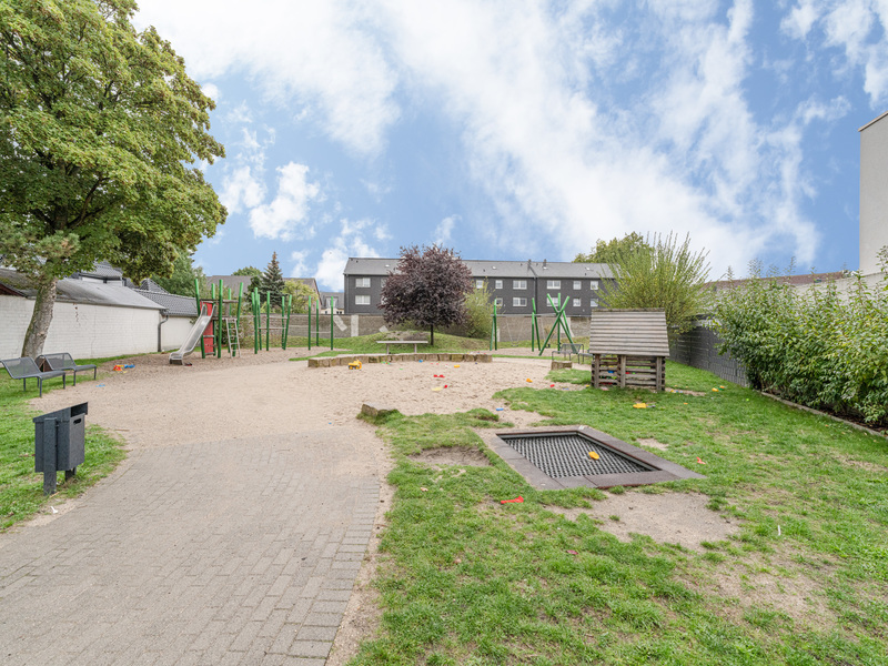 Spielplatz mit Sandfläche, Rutsche, Klettergerüst, Trampolin und Sitzbänken im Freien.