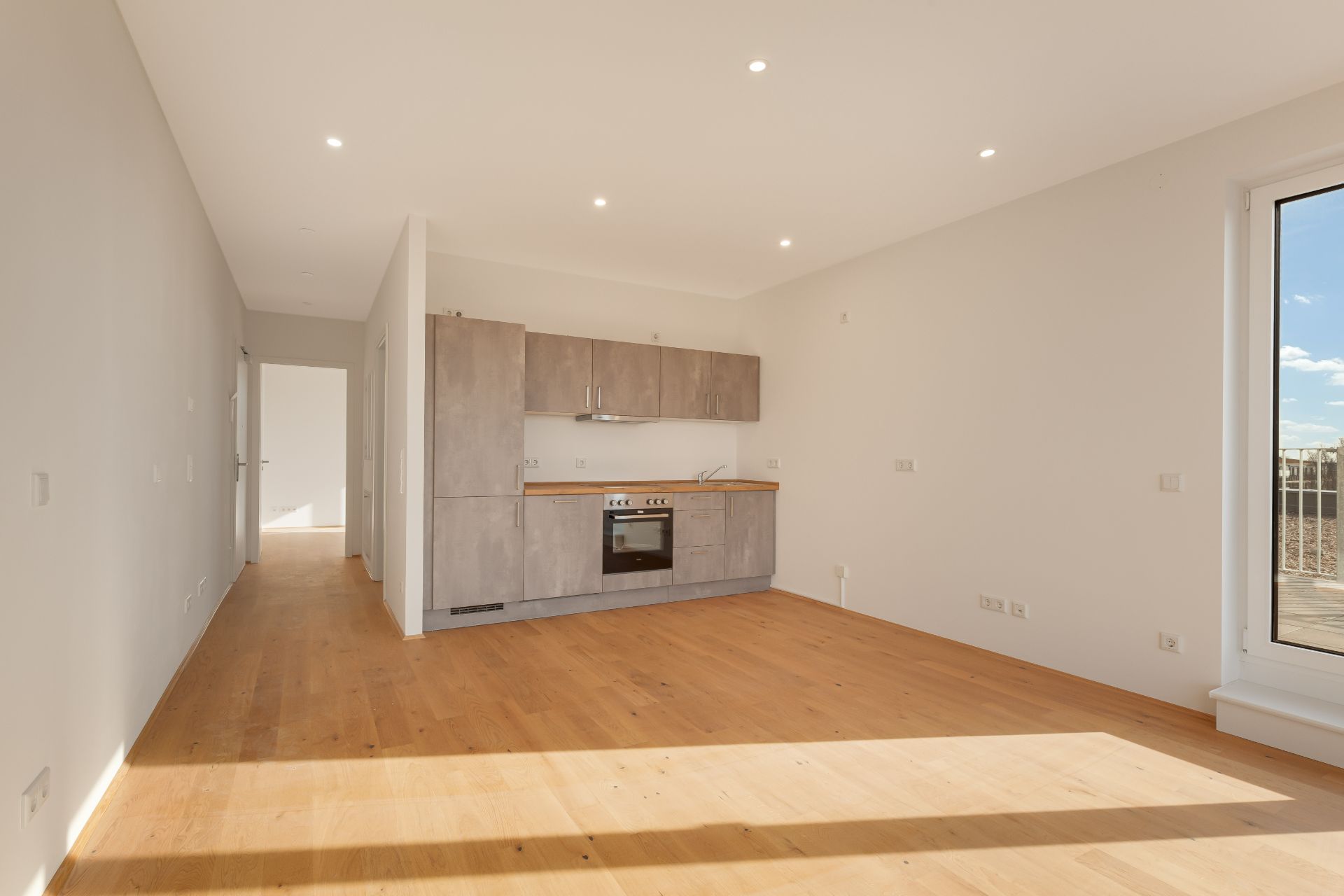 Helles Wohnzimmer mit Einbauküche, Holzfußboden und großer Fensterfront.
